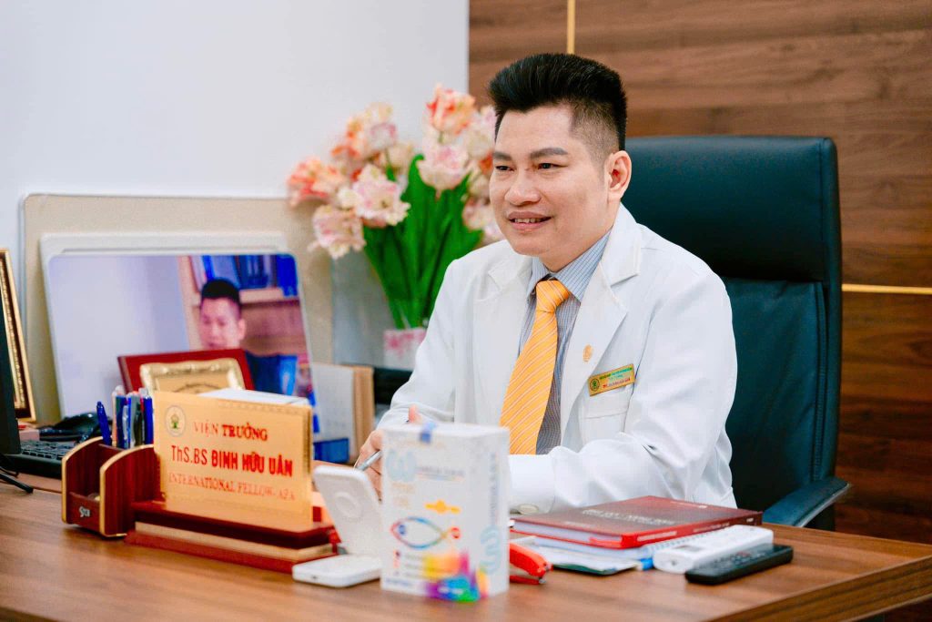 Bác sĩ chuyên khoa tâm thần Đinh Hữu Uân đang ngồi làm việc tại phòng khám chuyên khoa Tâm thần BS Uân, tay cầm bút và nở nụ cười thân thiện.